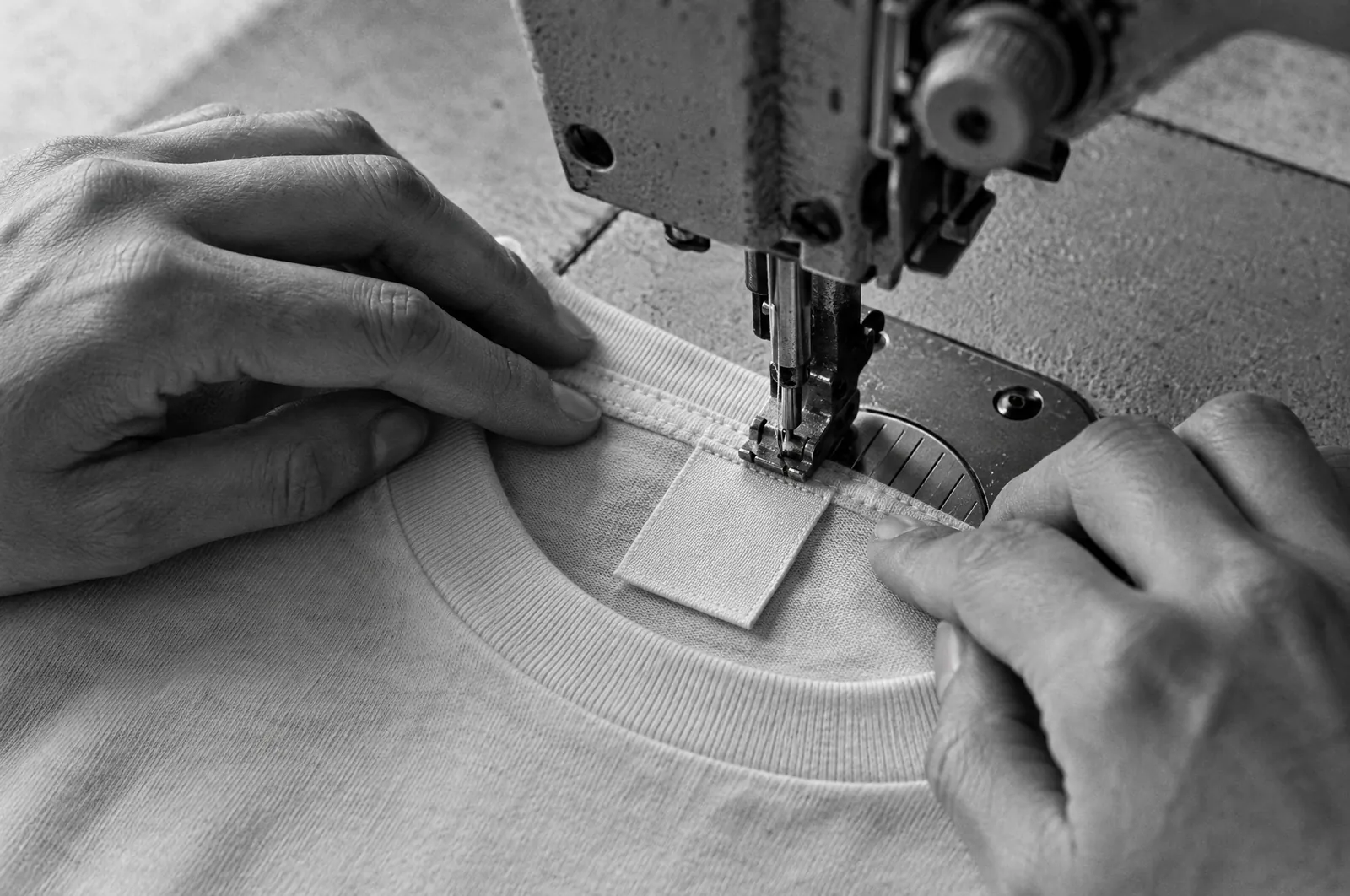 Black and white close-up photo of a sewing machine stitching a blank woven label into the inside neck of a plain T-shirt, with hands guiding the fabric.