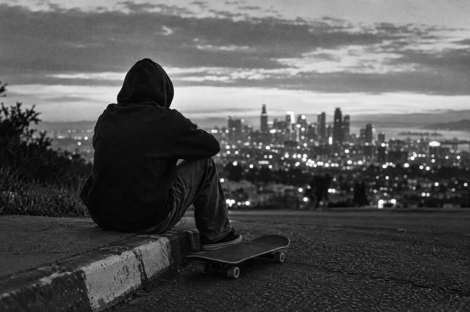Black and white photo of a skateboarder in a hood seen from behind, sitting on a painted street curb at dusk with a plain skateboard at their feet and city lights in the distance.