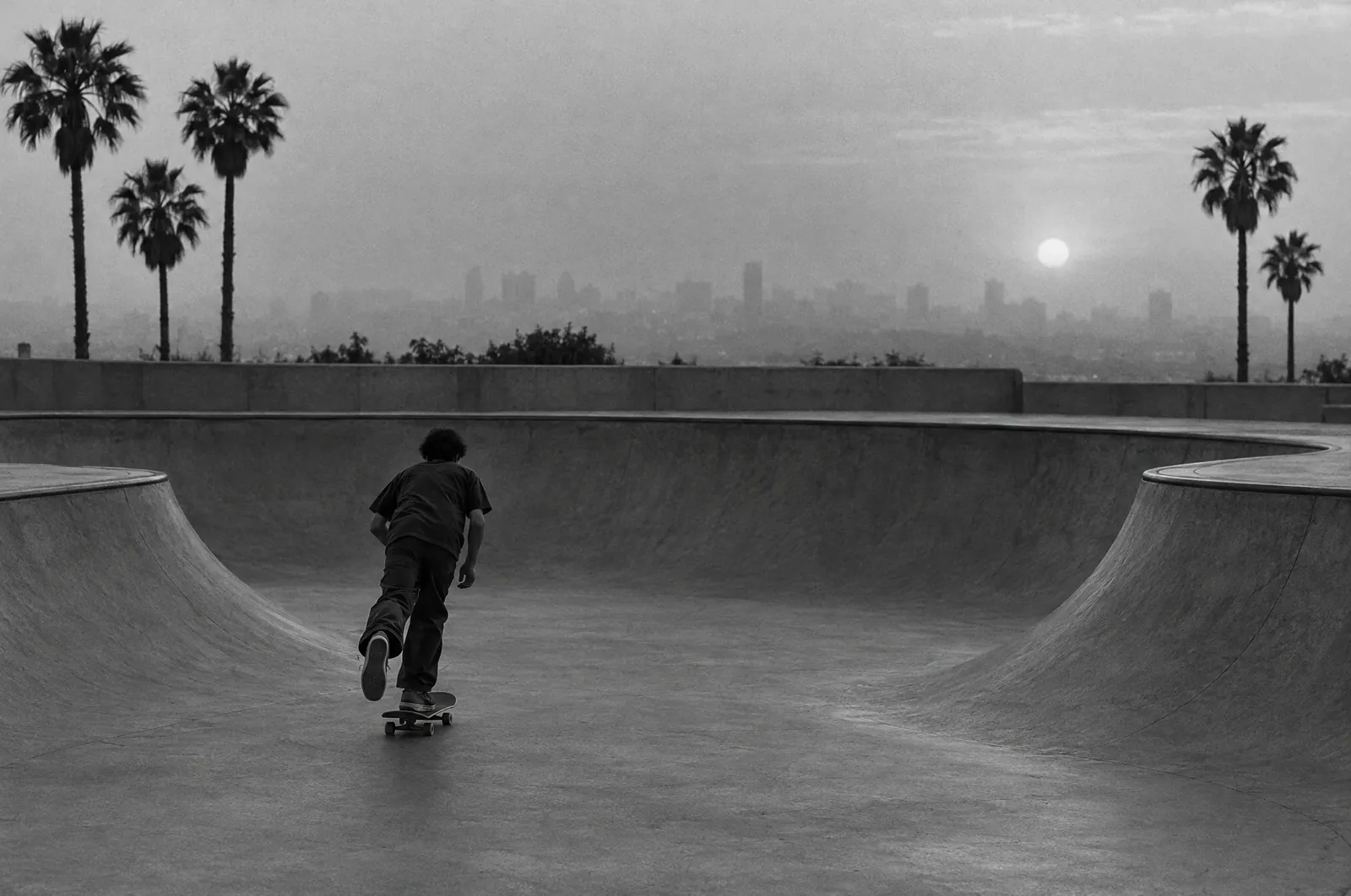 Black and white photo of a concrete skatepark bowl at dusk with a lone skater mid-push seen from behind, palm trees and a hazy city skyline behind.